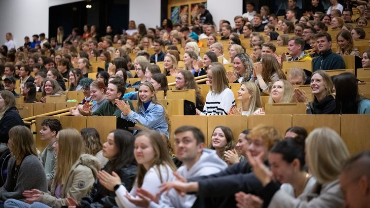 The picture shows a full hall. Students are sitting on the benches and in front of the stage.