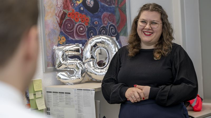 Das Bild zeigt Waleria Nichelmann in ihrem Büro. In der Bildmitte steht eine silberne 50. Nichelmann lächelt, während sie sich mit dem Interviewer unterhält.