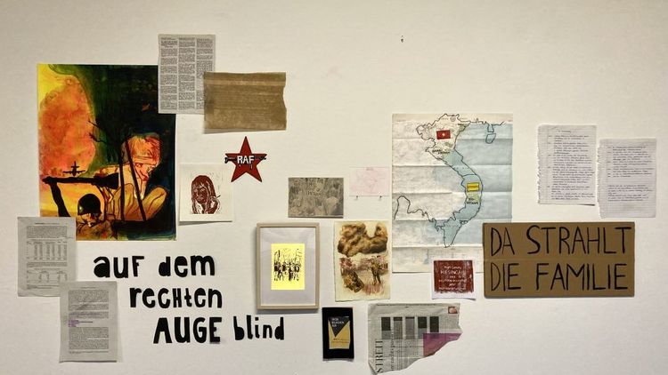 Das Bild zeigt eine Wand, auf der unter anderem der Slogan "auf dem rechten Auge blind", eine Karte von Vietnam, ein RAF-Sticker, Bilder von Kampfhandlungen und Ausschnitte aus Zeitungen angebracht sind.